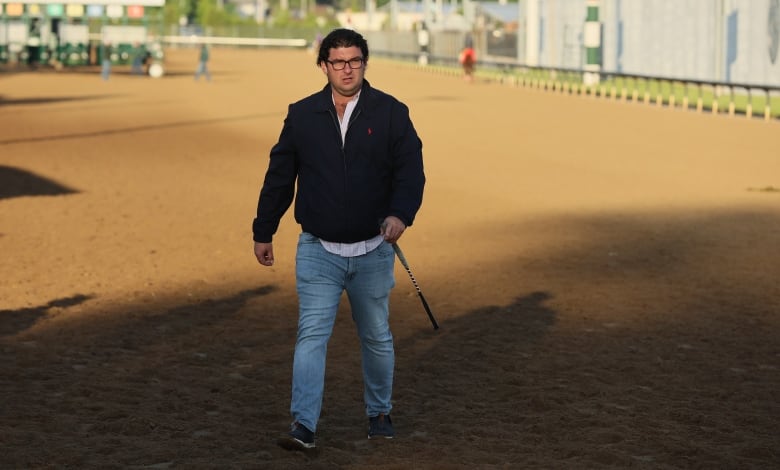 A bespectacled man in a jacket and jeans is shown walking on a dirt horse racing track.