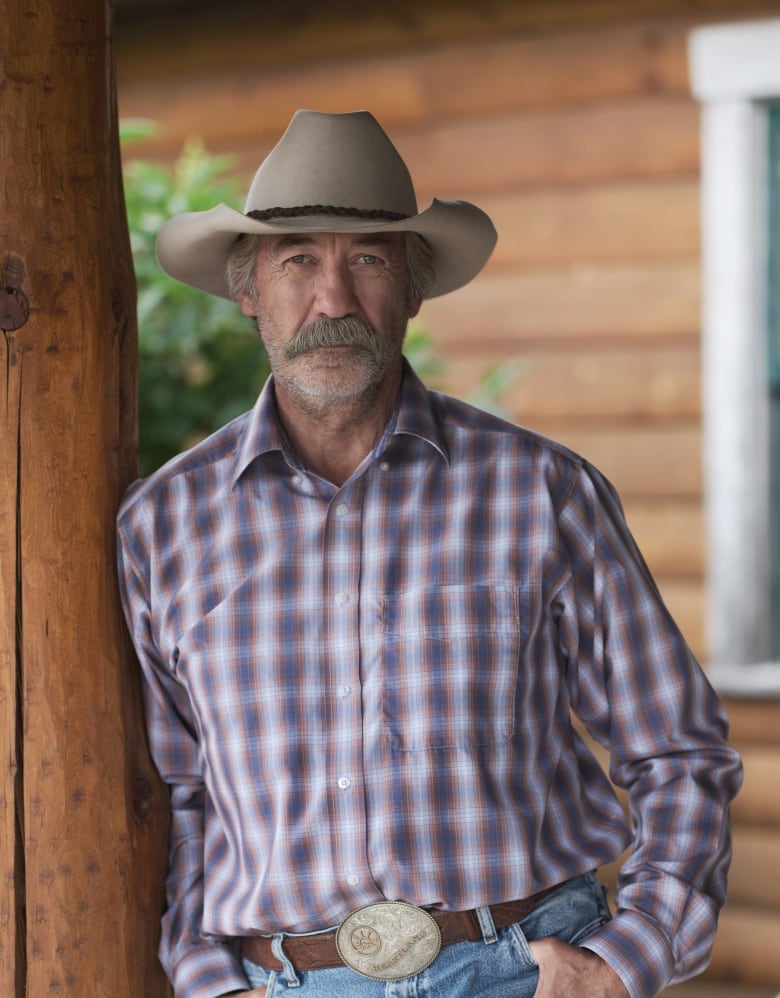 Actor Shaun Johnston in a purple and white plaid shirt and a tan cowboy hat, posing in front of a log-style ranch house.