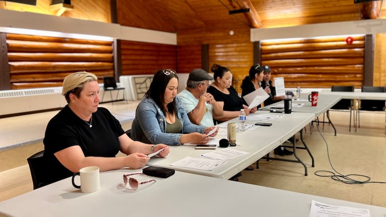 A row of six people sit at tables with papers in front of them.