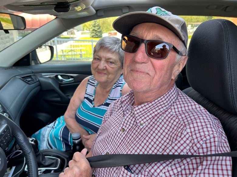 A man and woman sit in a car, looking out the driver's window. 