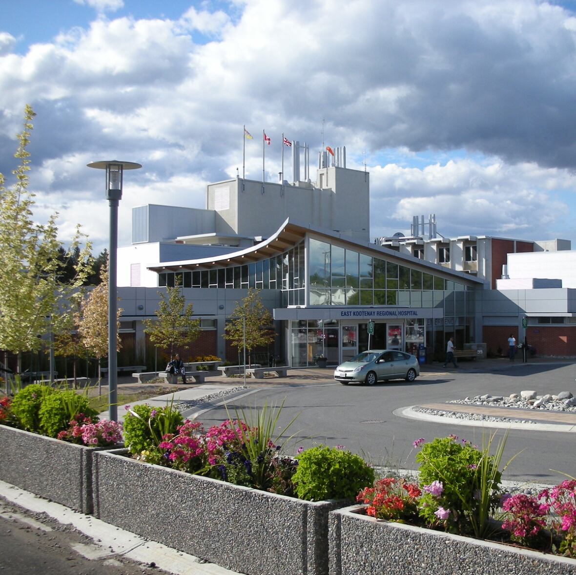 A silver car is parked outside the front doors of a grey hospital on a sunny day. A person is visible sitting at a picnic table.