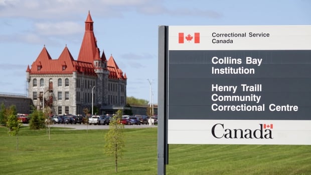 A grey sign identifying the prison behind it, a stone building that looks like a castle and has a red roof.