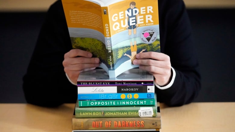 A person, seen only from the neck down, holds open a copy of the book Gender Queer. In front of her on a desk are a stack of books: The Bluest Eye, Lolita, l8r,gr8rm The Opposite of Innocent, Lawn Boy, Out of Darkness, and Beyond Magenta.