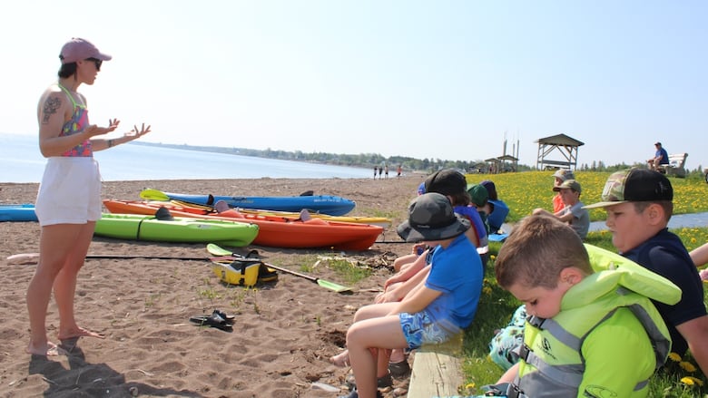 An Indigenous woman stands in front of kids on the beach. Some kids are wearing safety vests. Kayaks are laying on the beach. 