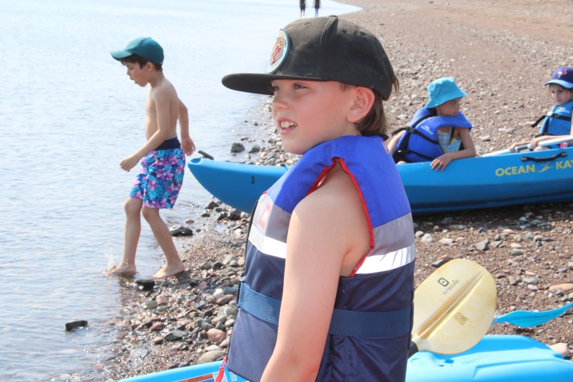 An Indigenous boy wears a life jacket near the beach, kids behind them are preparing to go in 