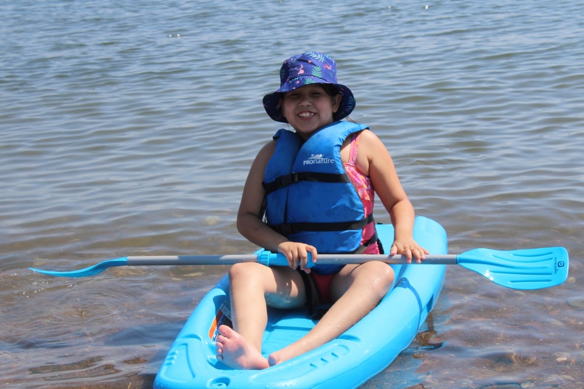 An Indigenous girl sits on a blue kayak with a blue paddle. Her kayak is in the water. 
