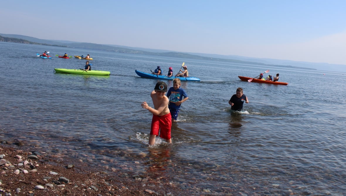 A group of Mi'kmaw boys are playing and running in the water. Six kayaks  coloured blue, green and red are in the background. 
