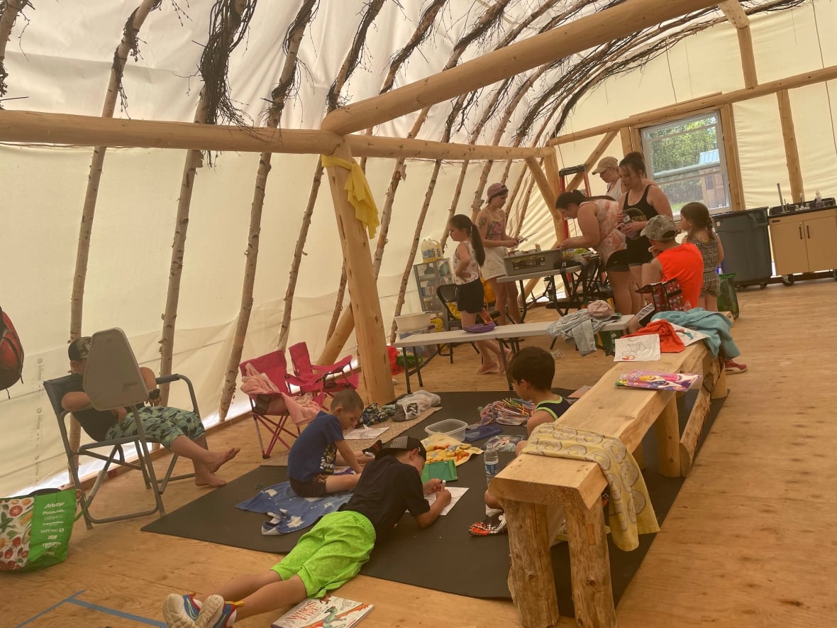 Indigenous students gather in a lodge with birch poles and tarp. Some kids lay on the floor while others line up for lunch.