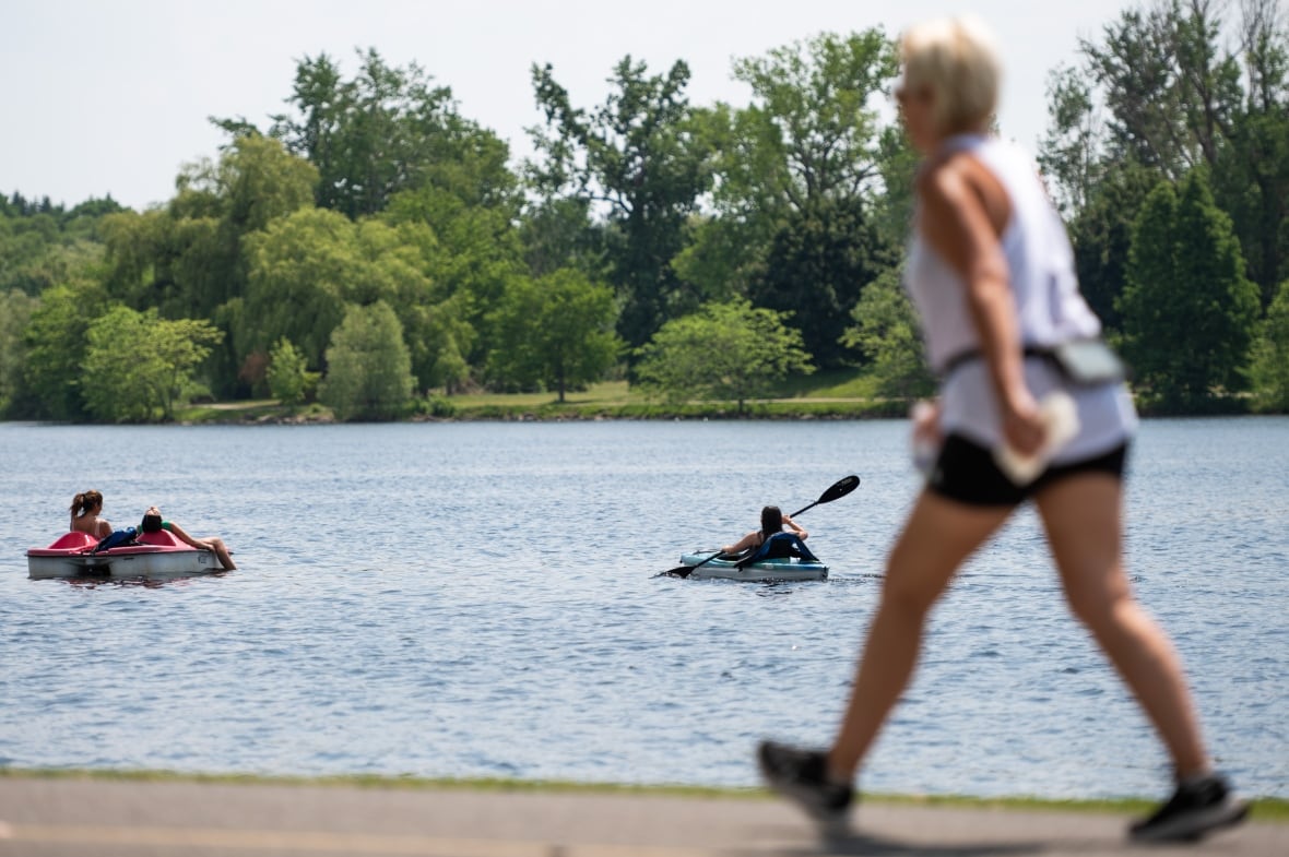 Someone walks next to a lake on a hot day. The lake has a kayaker and paddleboater on it.
