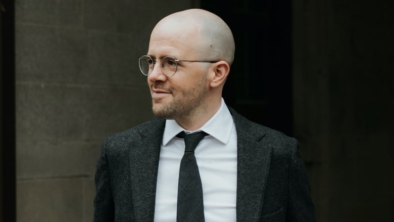 A bald man with round spectacles stands smiling in profile against a brick wall. He's wearing a black suit with a white shirt and black necktie.