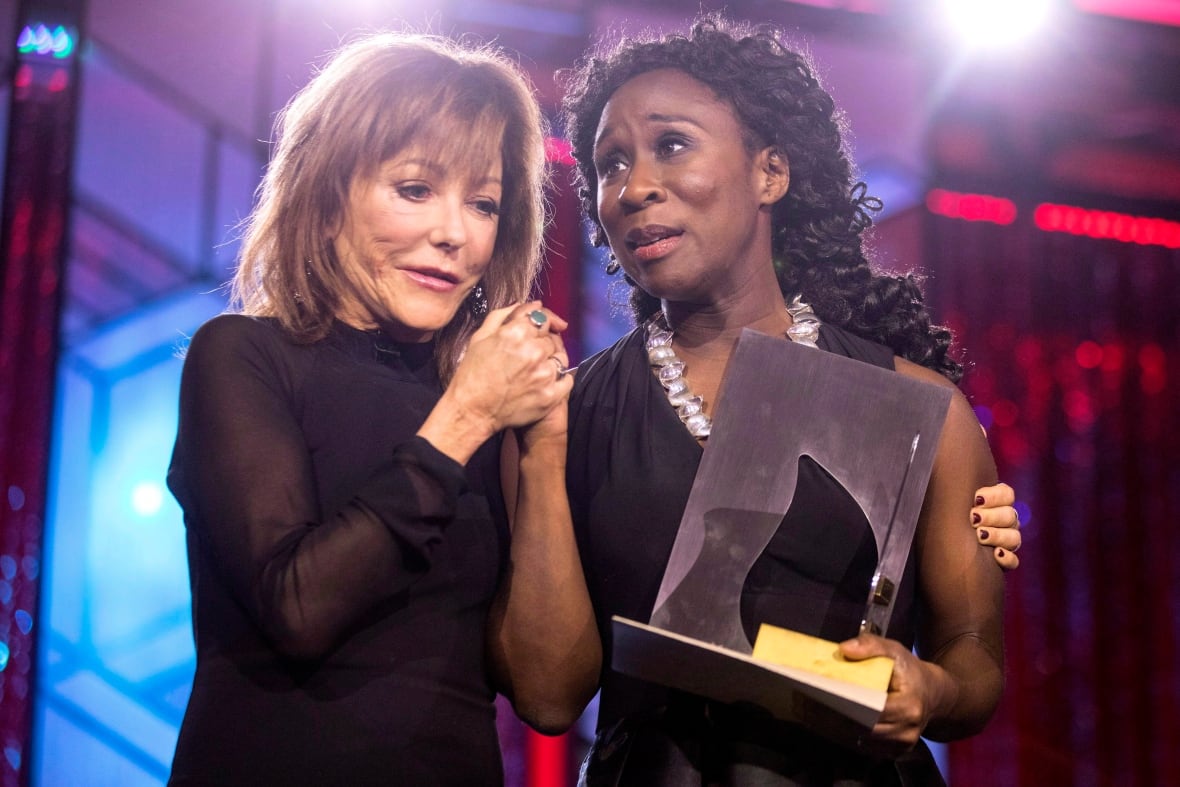 Two women in formal wear stand on stage. The white woman is holding the Black woman's hand and the Black woman is holding a trophy. Both look overwhelmed.
