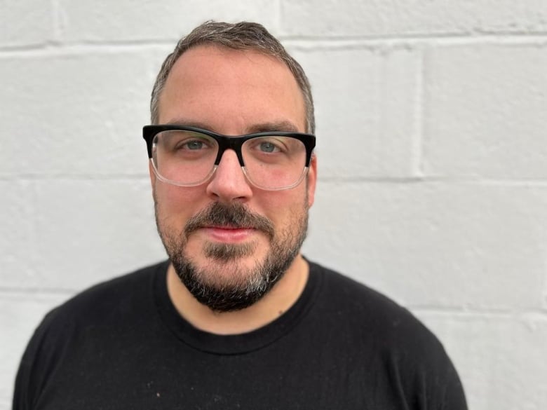 A man wearing glasses and a black shirt stands in front of a white brick wall.