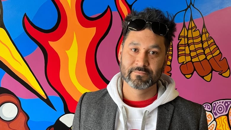 A man with a salt-and-pepper beard and a Detroit Red Wings jersey stands in front of a brightly-coloured mural on a wall. 