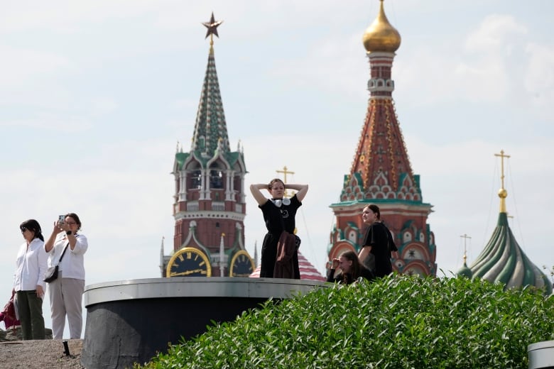 Five people are seen looking out from the perch of an urban hilltop park, with ornate buildings in the background. One woman stretches. One woman sits on a bench. And three others stand, with one taking a photo with her cellphone.