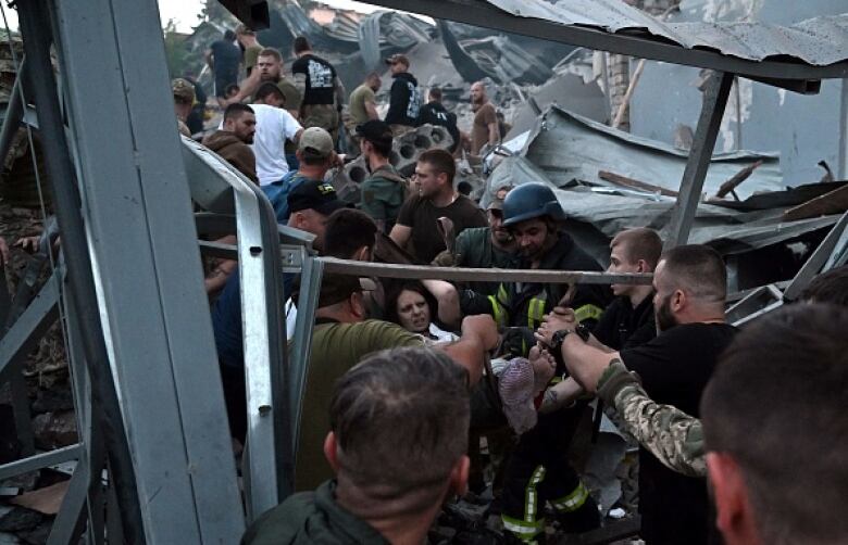 A woman is carried through a crowd amid a scene of damaged buildings and rubble.