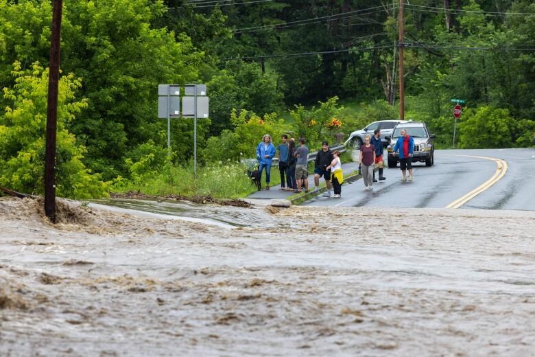 Water is shown flowing in the foreground of a photo as a group of people stand by the roadside in the background.