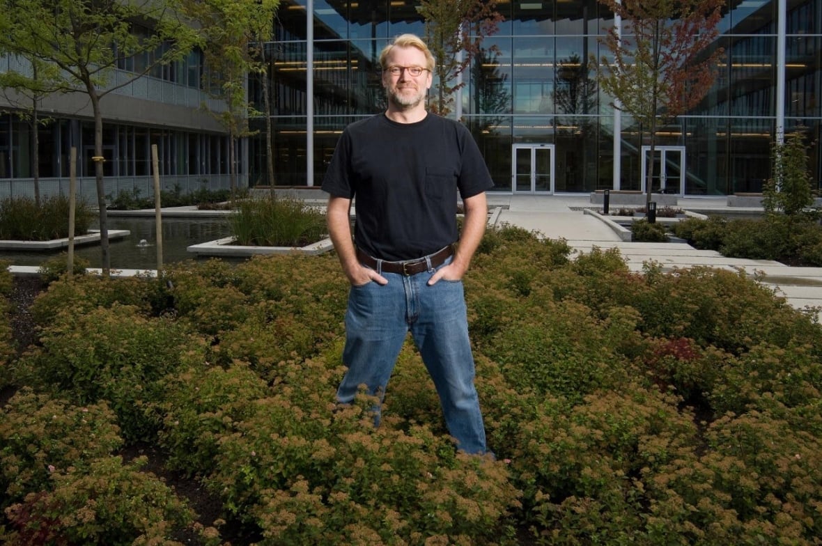A man in a dark t-shirt and jeans stands in bushes in front of a glass building and courtyard.