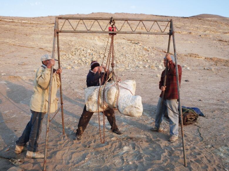 Three people with a huge white package hanging from a frame.