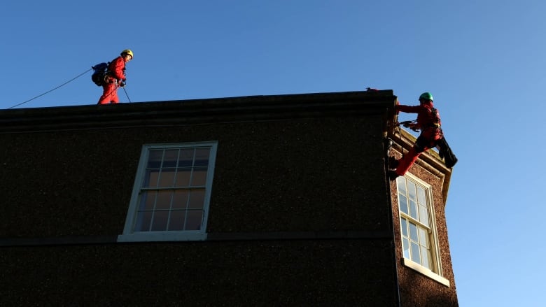 Two people in helmets and jumpers are shown scaling on the roof of a residential building.