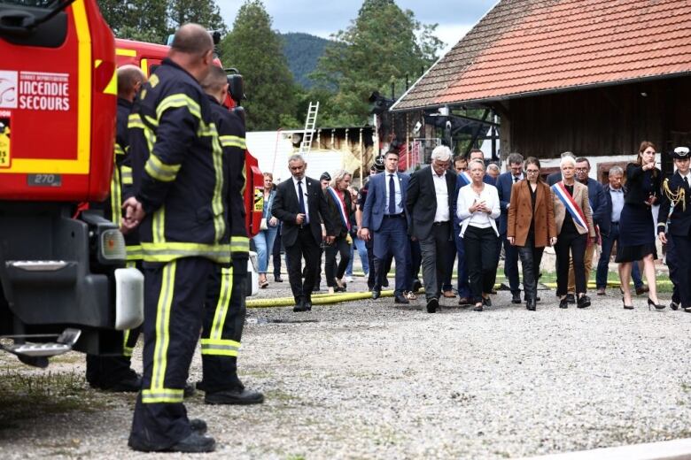 Government officials visit the scene of a fire as firefighters stand to the left.