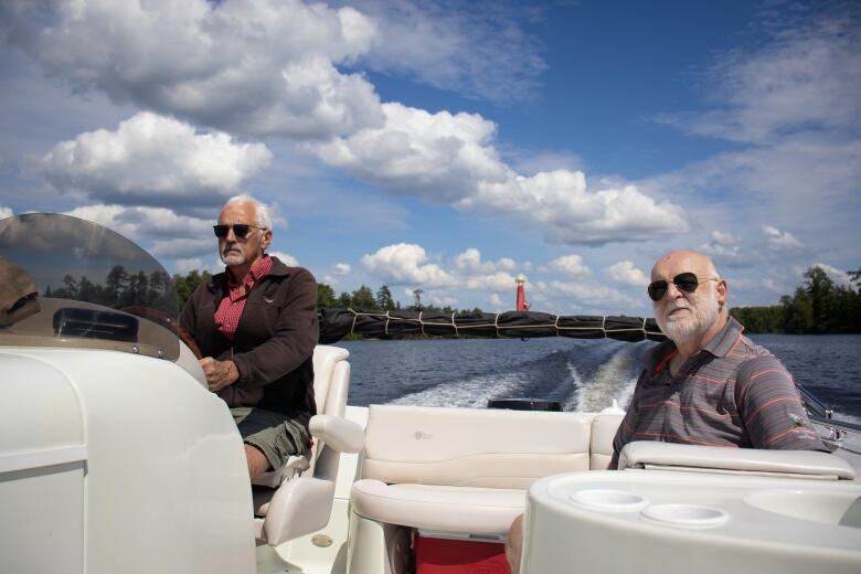 A man on the left steers a motorboat on a lake on a sunny day, while a second man on the right sits on the boat.