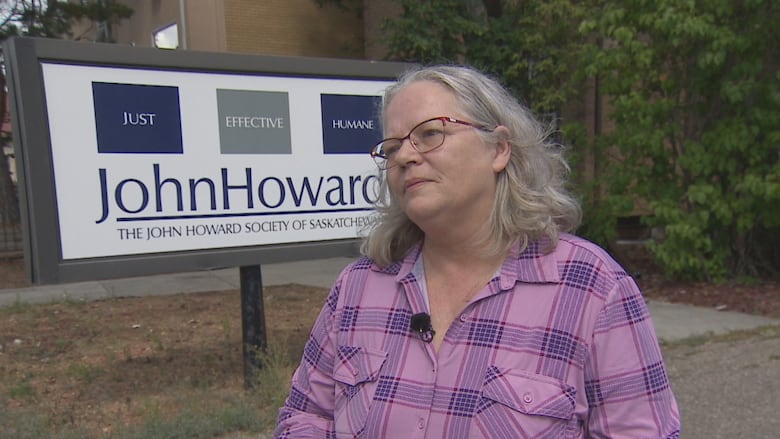 A woman with glasses and a purple button up shirt stands in front of the white, grey and blue John Howard Society of Saskatchewan sign. 