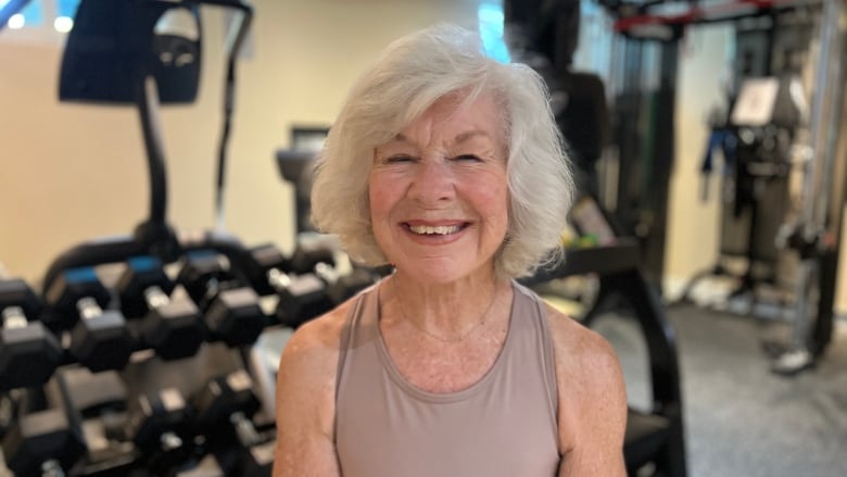 A smiling woman with white hair wears a tank top while standing in a gym. 