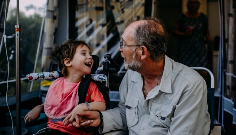 A little boy in a wheelchair smiles at an older man as they hold hands.