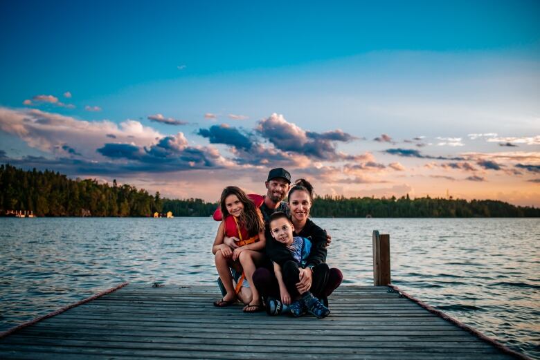 A young family is pictured on a dock on a lake around sunset, a mother and father are kneeling and holding a boy and girl.