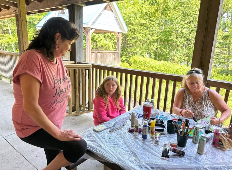 A woman wearing a peach-coloured shirt stands over a table where a woman and child sit. The table is covered in plastic tarp and painting supplies. 