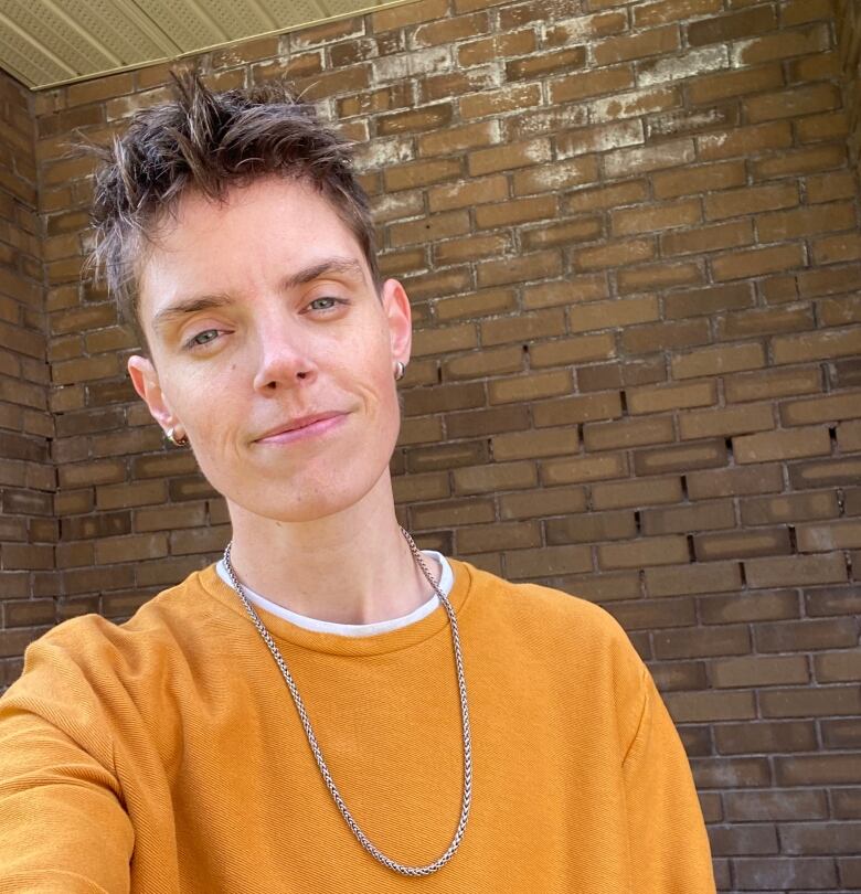 A person in a yellow sweater takes a selfie against a brick wall in the background.