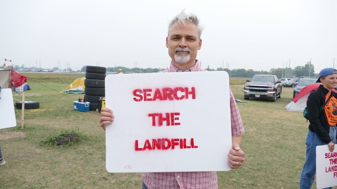 A man is standing outside with a sign that says "search the landfill."