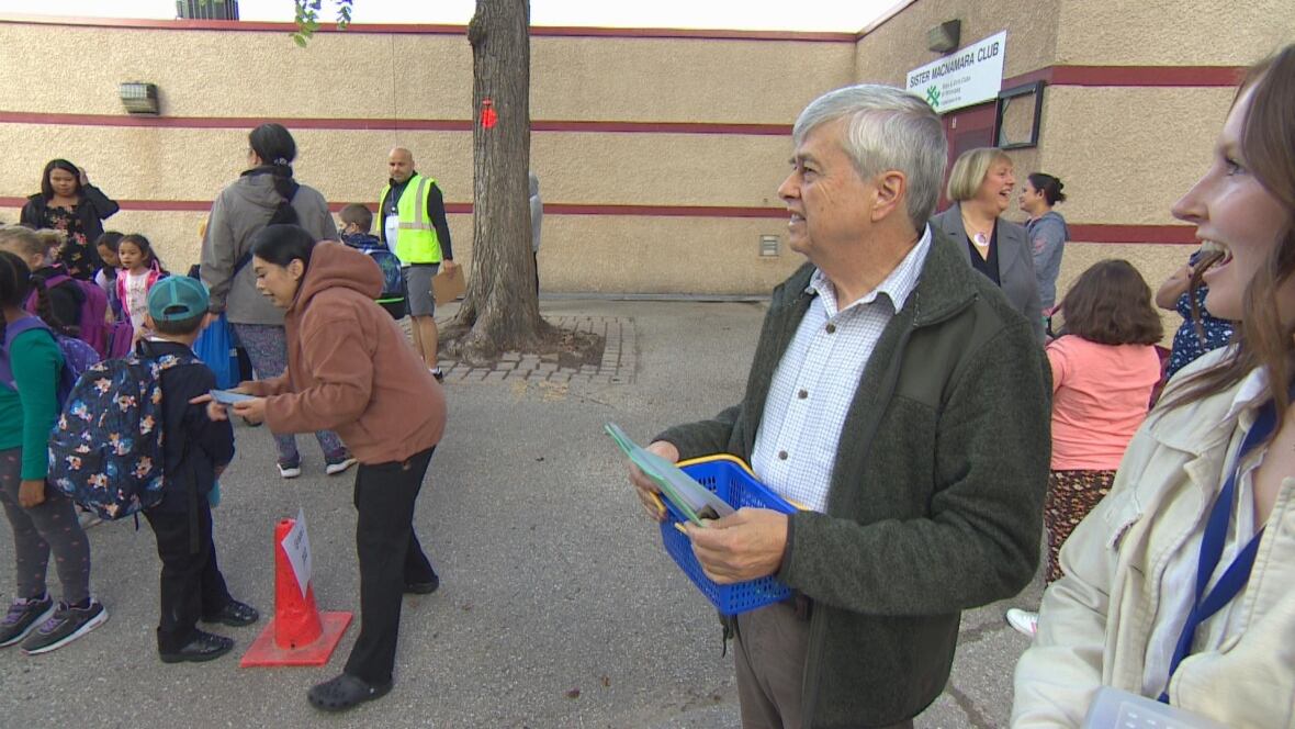 A teacher in a dark sweater and light button up shirt watches as his students arrive in a school yard. 