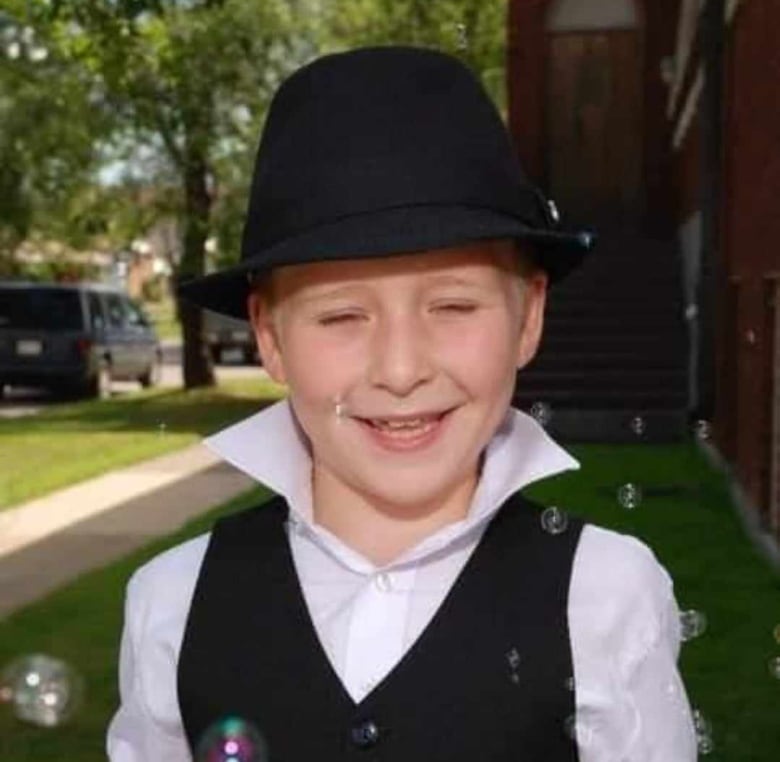 A childhood picture of Ashtyn, he is wearing a white coloured shirt and black vest with a black top hat and is smiling into the camera with bubbles around him.