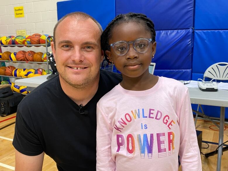 A man smiles as he crouches down next to a girl wearing glasses and a shirt that says 