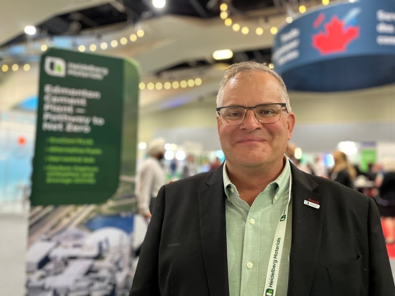 A man with glasses, a black jacket and green shirt smiles in front of a poster display for Heidelberg Materials at the Edmonton Convention Centre.