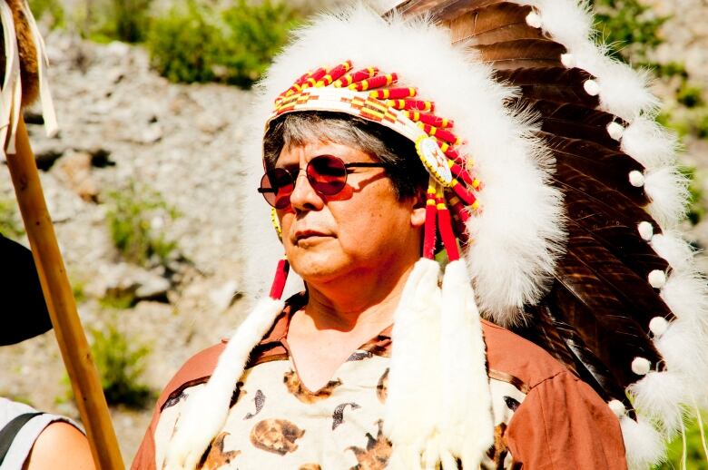 An Indigenous man wearing a large traditional headdress with red beads and black and white feathers stands in the sunshine. 