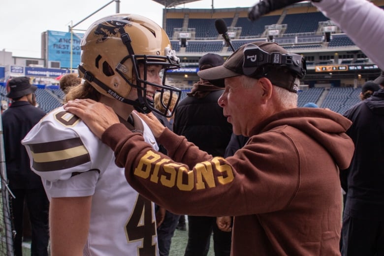 A football coach has his hands in the shoulders of a female football kicker, who smiles as they speak.