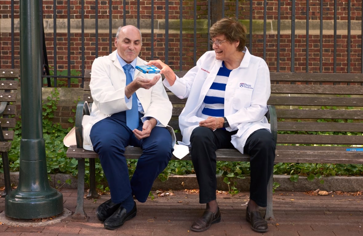 Dr. Weissman and Dr. Carico, on the same garden seat outside the building. She gives him something from a small box in his hand. It looks very fun while laughing.