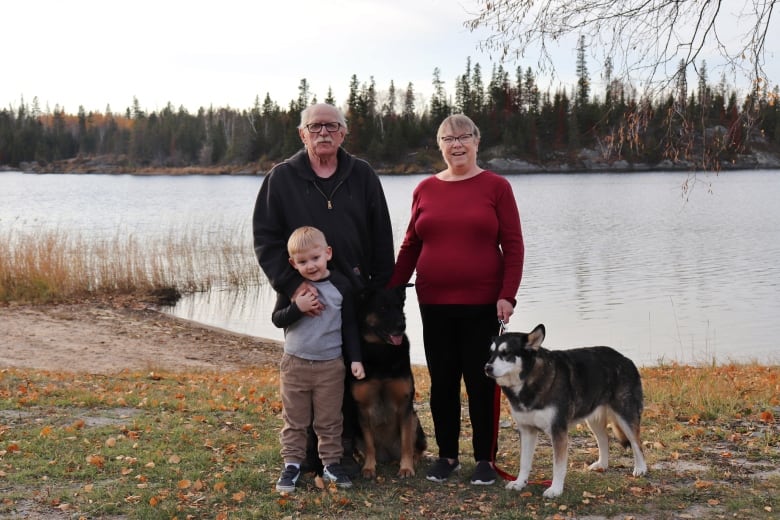A woman in a red shirt holds hands with a man wearing glasses. His arm is around a young boy, who is standing beside two dogs.