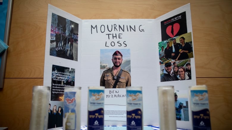 Six tall candles in a line in front of a three-sided board affixed with various photos of a young man.