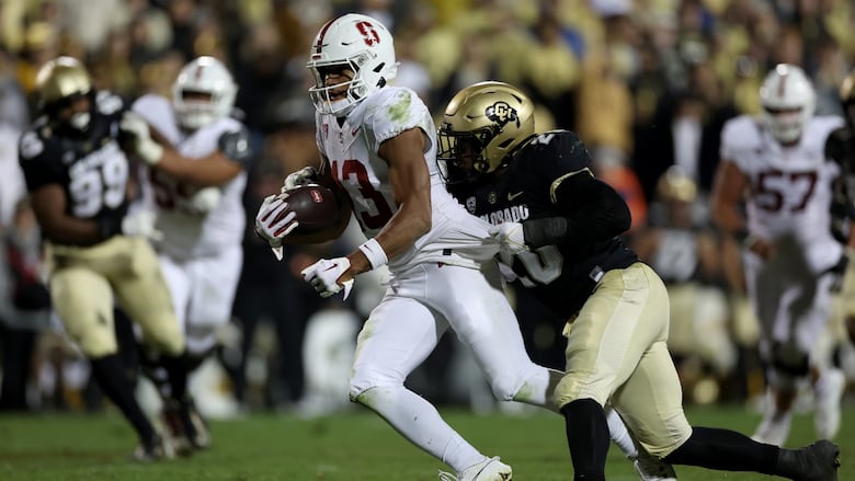 A football player in a white jersey and pants holds the ball while a player in a black and gold jersey attempts a tackle.