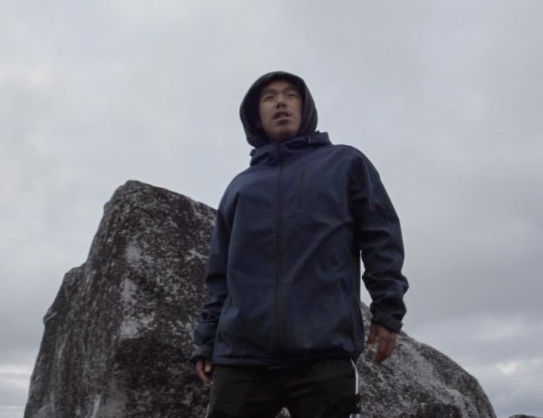 A young man stands near a large rock.