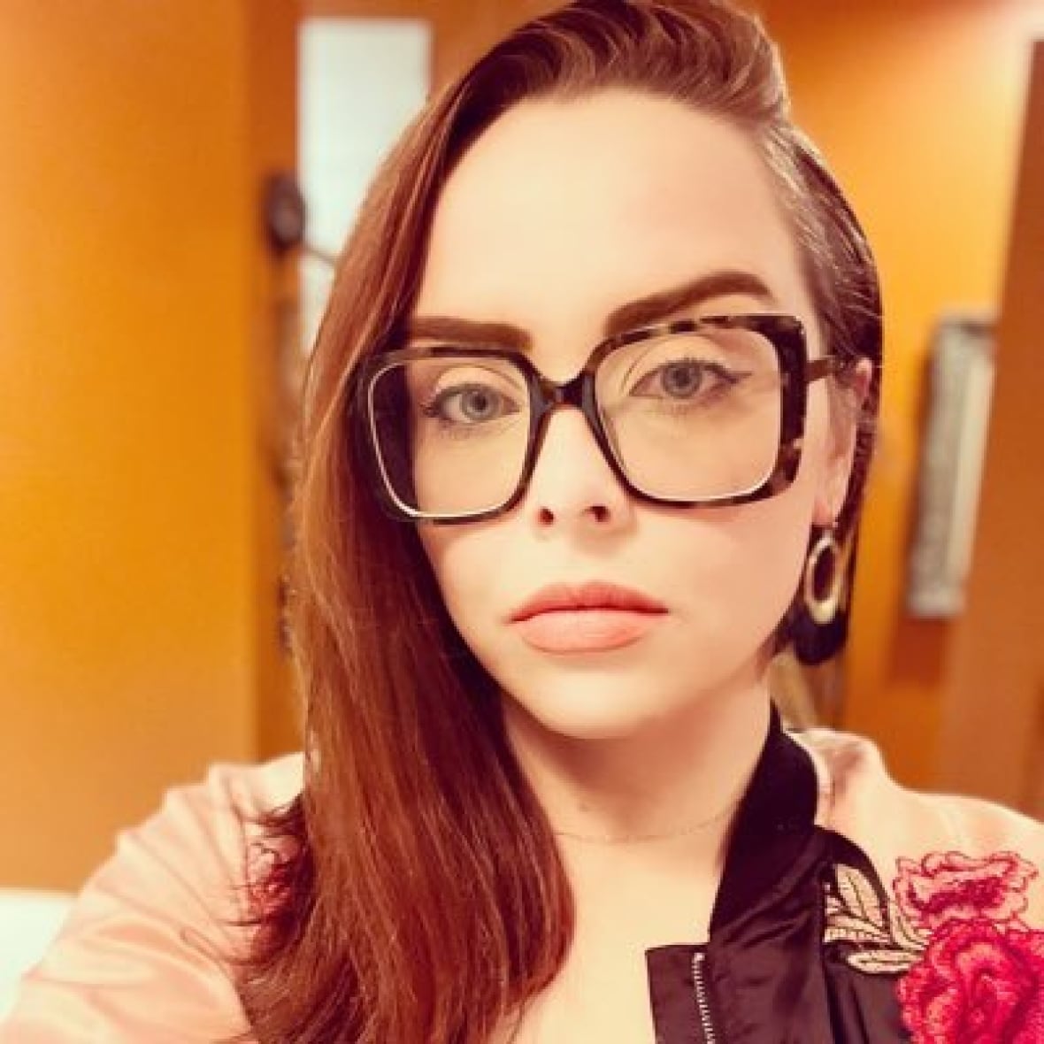 A woman posing in an orange-toned selfie. She is white with long brown hair and large, dark-rimmed glasses.