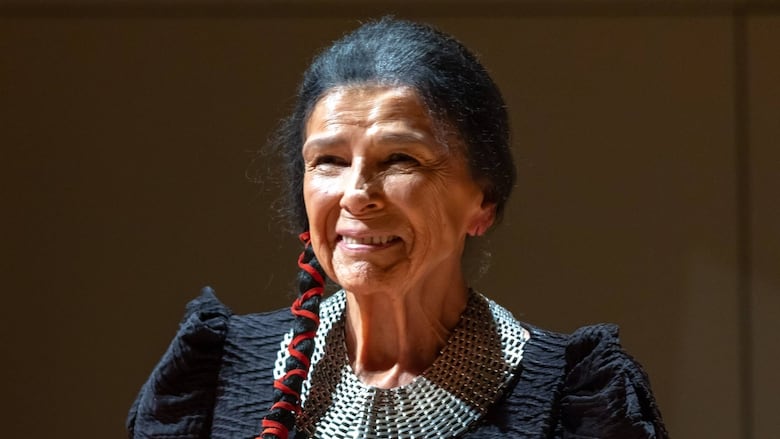 An Indigenous woman with long black hair tied up with red string is speaking at a podium.