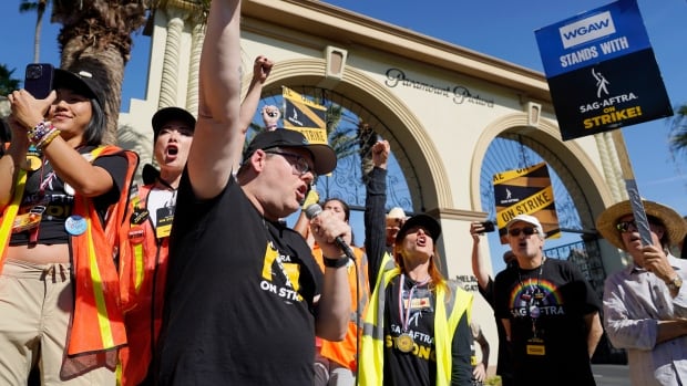 A group of chearing people wearing high-visibility vests stand outside of an arch adorned with the words "Paramount Pictures." A man standing close to the camera speaks into a microphone with his fist raised. A sign held by a protestor to his left reads "SAG-AFTRA STRIKE"