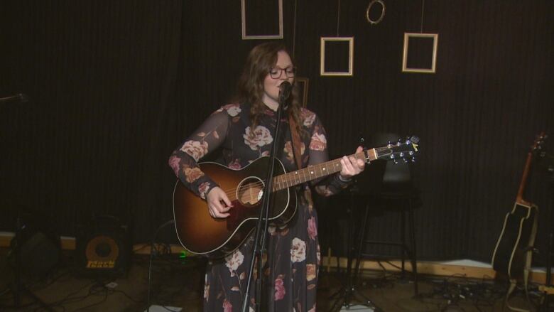 A woman in a black floral dress stands on a stage singing with a guitar. 