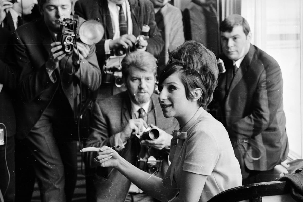 Black and white photo. Streisand sits, pointing her finger, as photographers surround her.