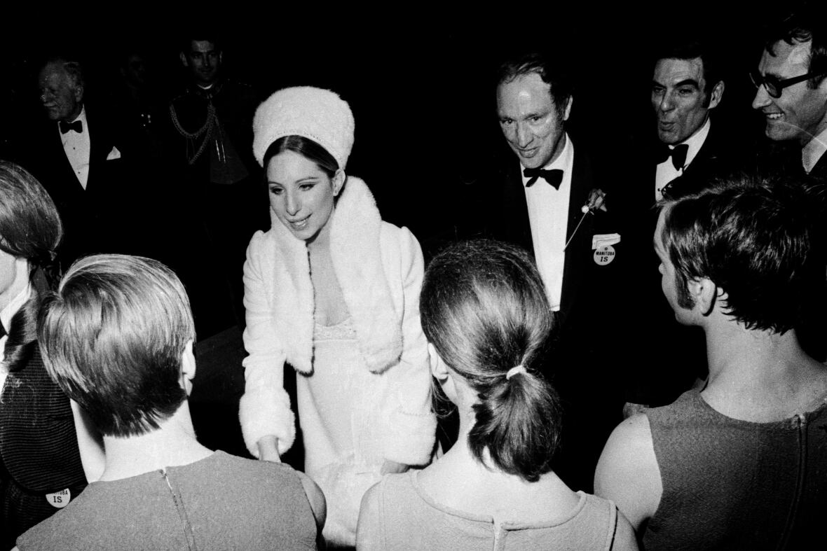 Black-and-white photo. Streisand and Pierre Trudeau walk through a crowd of people.