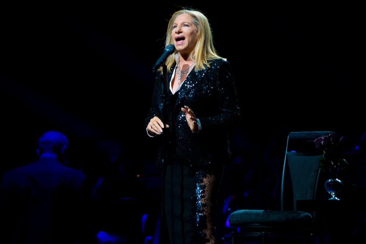 Streisand, wearing a long black sequined dress, performs behind a microphone on stage.